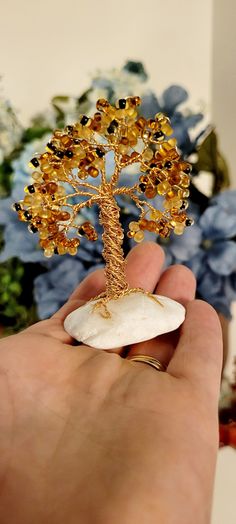 a hand holding a small tree made out of rock and glass beads in front of blue flowers