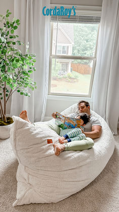 a man sitting on a bean bag chair reading a book to his child in front of a window