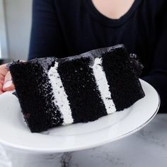 a woman holding a plate with a slice of black cake on it and white frosting