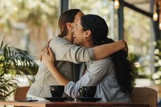 two women hugging each other at a table with coffee cups and plants in the background