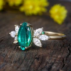 an emerald and diamond ring sitting on top of a piece of wood with flowers in the background