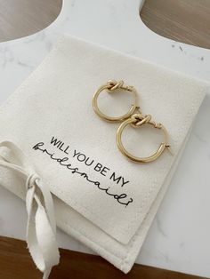 a pair of gold hoop earrings sitting on top of a piece of white paper with the words will you be my bridesmaid written on it