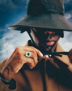 a man wearing a hat and holding scissors to his face with the sky in the background