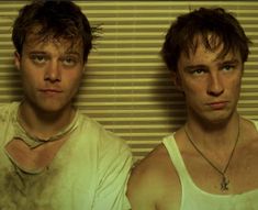 two young men standing next to each other in front of a wall with vertical blinds