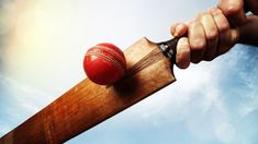 a hand holding a cricket bat with a red ball on the tip of it, in front of a blue sky