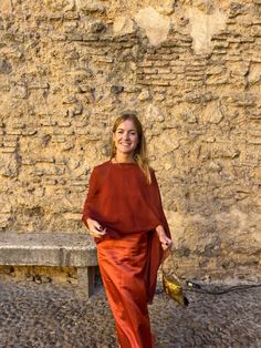 a woman standing in front of a stone wall wearing an orange dress and carrying a gold purse