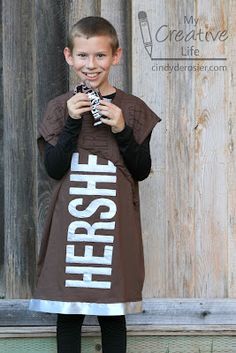 a young boy standing in front of a wooden wall holding a cell phone and wearing a brown shirt with the word hero printed on it