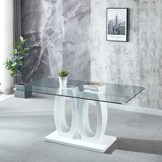 a glass table with two white legs and a plant on it in front of a gray wall