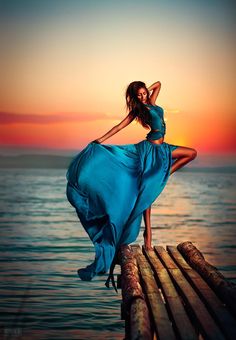 a woman in a blue dress is standing on a dock by the water at sunset