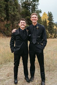 two men in black suits standing next to each other on a grass covered field with trees in the background