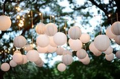 white paper lanterns hanging from strings in front of trees with string lights strung across them