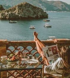 a woman sitting at a table reading a magazine overlooking the ocean and boats in the water