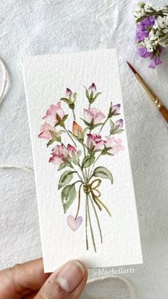 a hand holding a card with watercolor flowers on it next to some scissors and thread