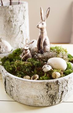 two rabbits sitting in a bowl filled with moss and eggs on top of a table