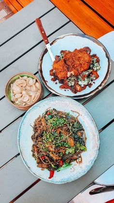 two plates of food sitting on top of a wooden table next to bowls of noodles