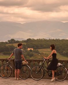 a man and woman standing next to their bikes looking at each other with mountains in the background