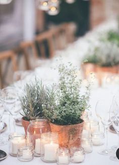 a table topped with lots of candles and vases filled with greenery next to wine glasses