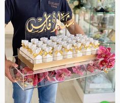 a man is holding a tray with many small cakes on it and flowers in front of him