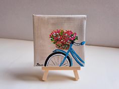 a small canvas with a blue bicycle and flowers on it is sitting on a wooden easel