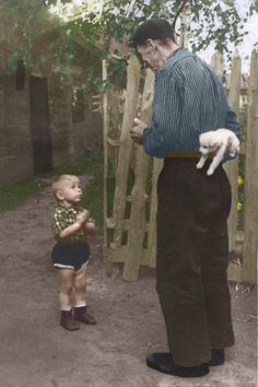 an old man standing next to a little boy in front of a fence with a cat on his arm