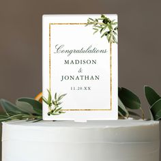 a white cake with greenery on top and an acrylic sign that reads congratulations madison & jonathan