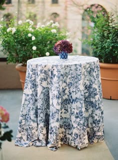 a blue and white table cloth on top of a round table in front of potted plants