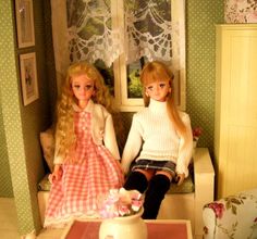 two dolls are sitting on a couch in a doll house, one is wearing a pink dress and the other wears a white sweater