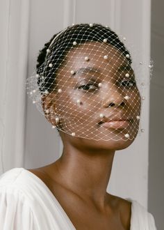 a woman wearing a birdcage veil with pearls on her head and shoulders, in front of a white wall