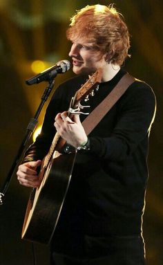 a young man with red hair is singing into a microphone and holding an acoustic guitar