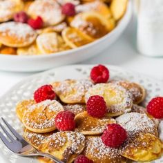 a white plate topped with pancakes covered in powdered sugar and raspberries next to a bowl of fruit