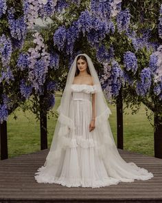 a woman wearing a wedding dress and veil standing in front of purple flowers on a wooden deck