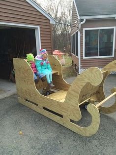 two children are sitting in a wooden sleigh on the driveway next to their garage