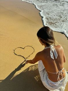 a woman sitting on the beach drawing a heart in the sand