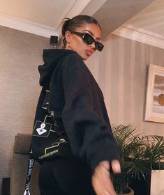 a woman wearing sunglasses and a black hoodie is standing in a room with a potted plant