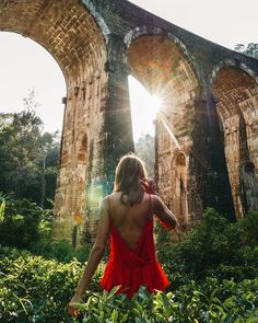 a woman in a red dress is looking at the sun through an old stone bridge