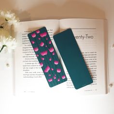 two bookmarks sitting on top of an open book next to a vase with flowers