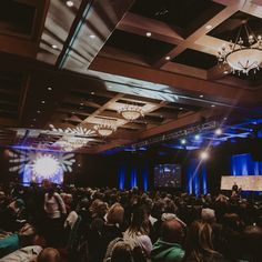 a large room filled with people sitting at tables and standing in front of a projection screen