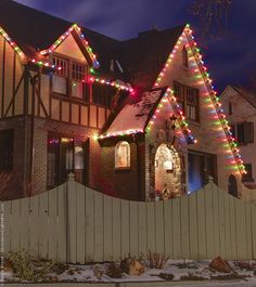 a large house with christmas lights on it's roof and windows in front of a white picket fence