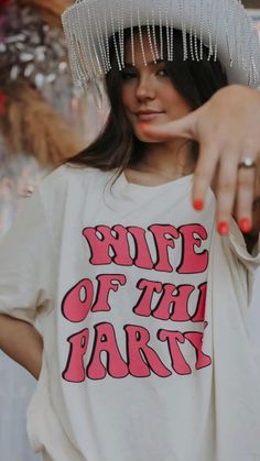 a woman pointing at the camera wearing a white shirt with pink letters on it and fringe around her head