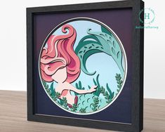 a painting of a mermaid with pink hair and green leaves in a black frame on a wooden table