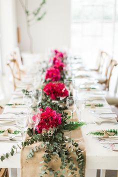 a long table with red flowers and greenery on the top, along with place settings