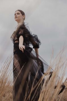 a woman standing in tall grass wearing a long black dress with feathers on her shoulders