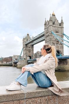 a woman sitting on the edge of a wall next to a body of water with a bridge in the background