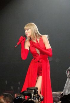 taylor swift performing on stage in red dress with microphone and man behind her looking off to the side