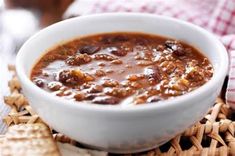 a white bowl filled with chili and cheese on top of a woven place mat next to crackers