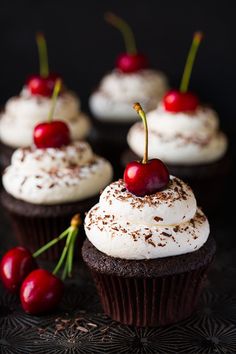 chocolate cupcakes with white frosting and cherries on top, sitting on a black surface