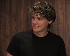 a young man with curly hair and black shirt smiling at someone in front of a wooden wall