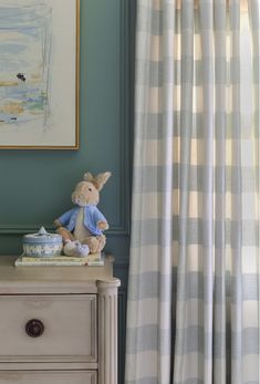 a teddy bear sitting on top of a dresser next to a painting and window curtain