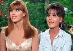 two women sitting next to each other in front of green plants and trees, one with blue eyes