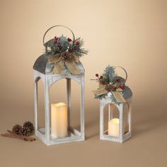 two white lanterns decorated with holly and pine cones are sitting next to each other on a brown background